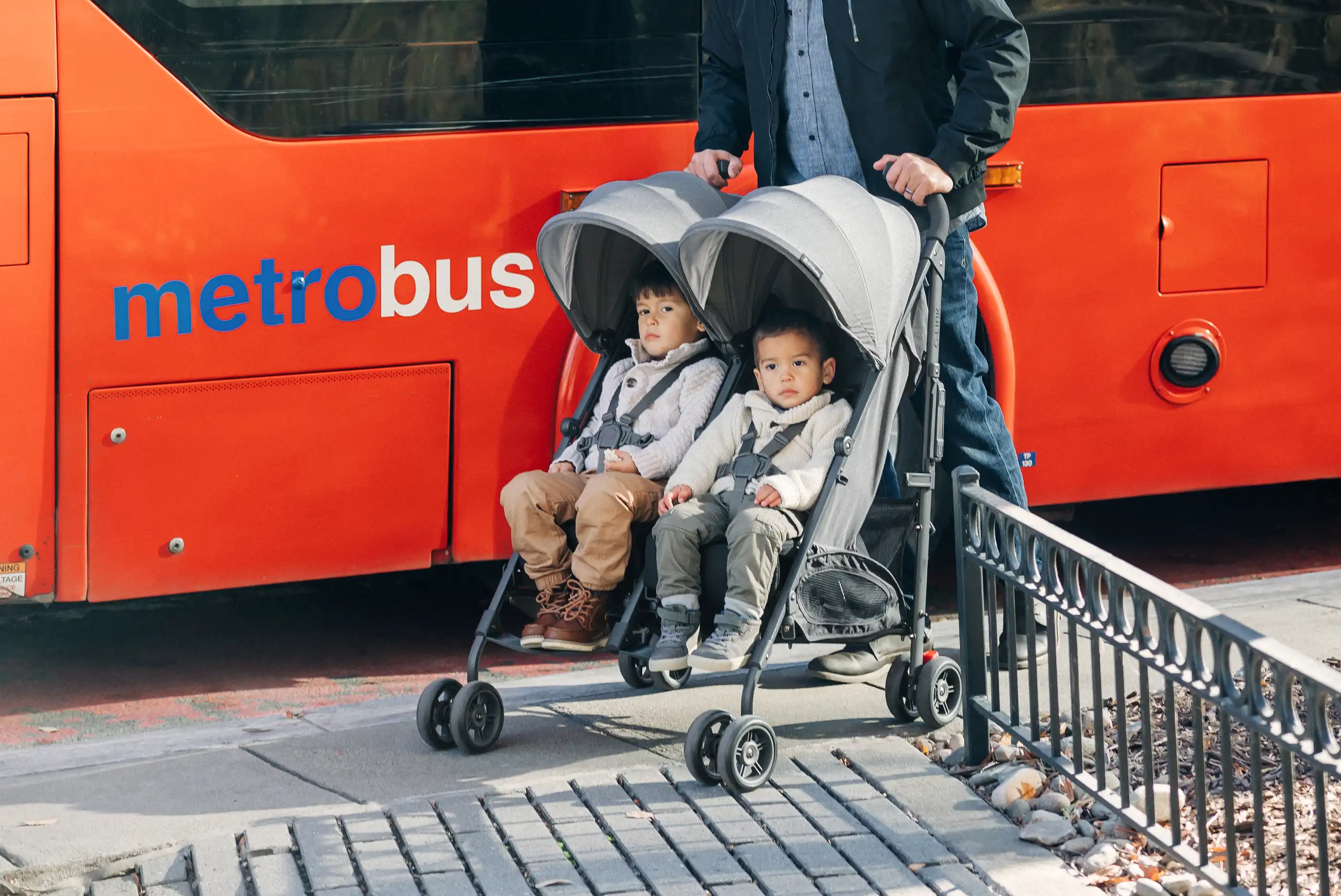 Parent pushing two children on a city sidewalk, next to a bus